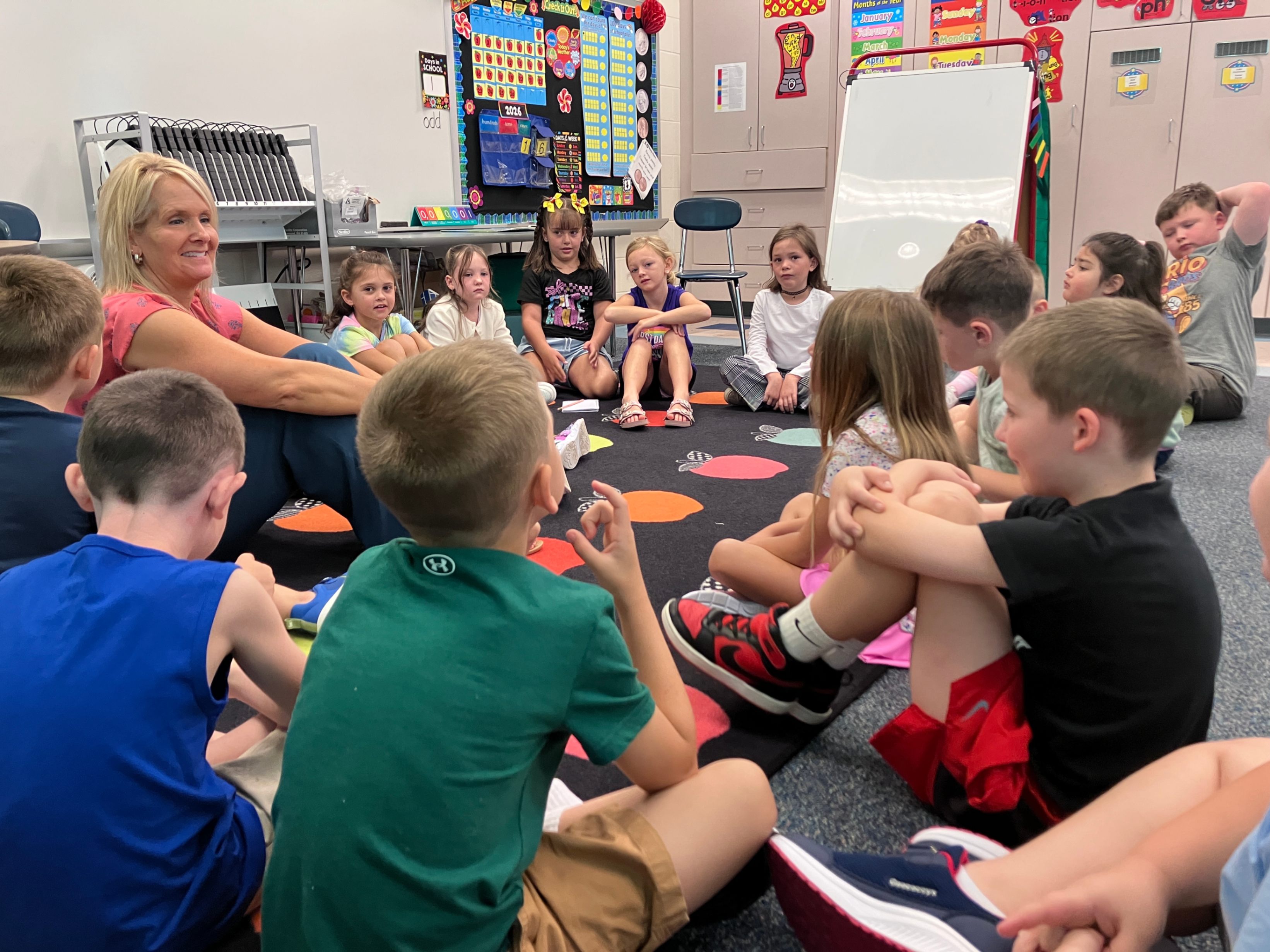 Mrs. Clinger and her first grade students sit on the floor together in their classroom.